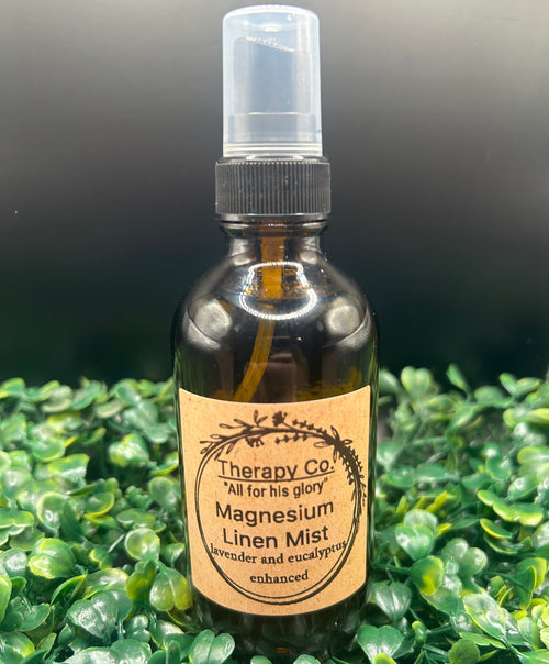 A dark amber glass spray bottle labeled 'Therapy Co. Magnesium Linen Mist' with a clear spray top, set against a background of green foliage.